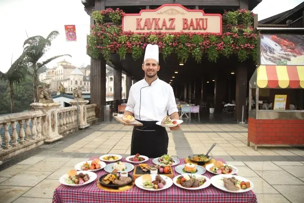 Chef présentant une variété de plats gastronomiques au restaurant Kavkaz Baku, situé dans un décor extérieur pittoresque avec une architecture traditionnelle et une verdure luxuriante, parfait pour dîner et voyager.