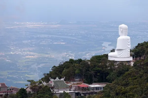 Grande statue de Bouddha blanc sur une colline surplombant une ville et une rivière, entourée d'une verdure luxuriante et de bâtiments traditionnels, idéale pour les voyages et le tourisme spirituel.